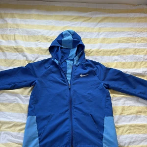 Nike blå vindjacka med huva - Nike vindjacka i två nyanser av blått med huva och dragkedja. Jackan har sportig look, fickor framtill och en snygg ljusblå detalj på huvan och ryggen. Perfekt för dig som gillar streetwear och vill ha något lätt och stilrent.