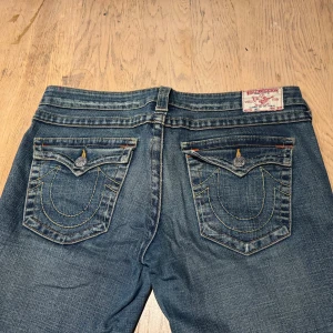 True Religion blå jeans med broderade fickor - Säljer ett par blå jeans från True Religion med ikoniska broderade bakfickor och snygga kontrastsömmar. Det är bootcut jeans med detaljerade knappar och etikett bak i midjan. Lite slitna längst ned. Annars fel fria. Lite långa på mig som är över 170. 