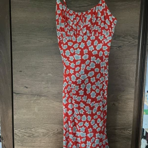 Röd blommig  med rosett - Säljer en röd klänning med vita och ljusblå blommor. Klänningen har smala axelband, öppen rygg med stor rosett och en figurnära passform. 