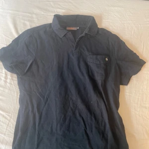 Mörkblå pikétröja från Morris - Snygg mörkblå pikétröja från Morris med klassisk krage och korta ärmar. Bröstficka med broderad logga i form av en lilja. Tillverkad i mjuk bomull som känns skön mot huden. Perfekt för en avslappnad men stilren look. Pris kan diskuteras. 