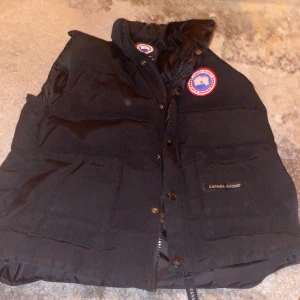 Svart dunväst från Canada Goose - Säljer en svart dunväst från Canada Goose med knappar och dragkedja. Västen har en hög krage och är perfekt för kyligare väder. Den ikoniska loggan syns tydligt på bröstet.