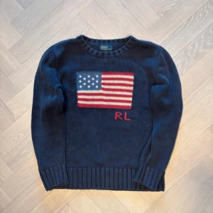 Mörkblå stickad tröja från Ralph Lauren - Säljer en snygg mörkblå stickad tröja från Ralph Lauren med en amerikansk flagga på framsidan. Tröjan har långa ärmar och ribbade kanter vid ärmslut och nederkant. Perfekt för kyliga dagar!