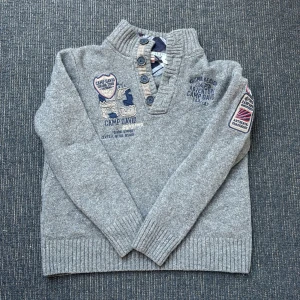 Grå stickad tröja - Säljer en snygg grå vintage tröja från Camp David med broderade detaljer och knappar framtill. Tröjan har en hög krage och långa ärmar, perfekt för kyligare dagar. Den är ribbad vid ärmslut och nederkant för en bekväm passform. Perfekt för skola osv. Storlek XL men sitter som L.