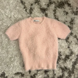 Rosa stickad tröja från Zara - Säljer en söt rosa stickad tröja från Zara. Den har korta ärmar och en mjuk, fluffig textur som ger en mysig känsla. Perfekt för att lägga till en touch av färg och stil till din garderob. Passar bra till både jeans och kjol!