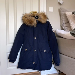 Mörkblå parkajacka med päls från Svea - Säljer en snygg mörkblå parkajacka från Svea med fuskpäls på huvan. Jackan har dragkedja och knappar framtill samt två stora fickor. Perfekt för kalla dagar!