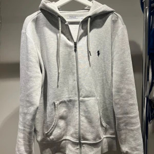 Grå hoodie från Ralph Lauren - Säljer en stilren grå hoodie från Ralph Lauren med dragkedja och justerbar huva. Tröjan har en liten broderad logga på bröstet och praktiska fickor framtill. Perfekt för en avslappnad look. Hör av er vid frågor!