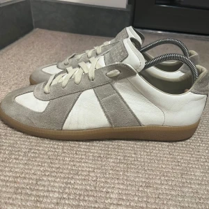 Maison Margiela sneakers i vitt och grått - Snygga sneakers från Maison Margiela i en stilren kombination av vitt och grått. Skorna har en klassisk design med snörning och en bekväm gummisula. Perfekta för en avslappnad och trendig look. Storlek 43. Nypris: 6800. Box ingår