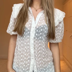 Vit blus från Ellos - Säljer en jättesöt vit blus från ellos. Blusen har puffärmar och en stor krage med volanger. Blusen är lätt genomskinlig och har blom detaljer. Tyvärr saknas en knapp, som tur är finns en extra knapp som lätt kan sys på!💖