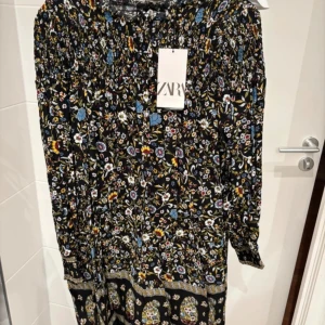 Blommig klänning från Zara - Säljer en färgglad blommig klänning från Zara med långa ärmar och ett detaljerat mönster. Klänningen har en elastisk midja och är perfekt för en bohemisk look. Den har en mörk bas med inslag av blått, gult och rött i mönstret.