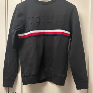 Svart sweatshirt från Tommy Hilfiger - Snygg svart sweatshirt från Tommy Hilfiger med ett upphöjt 'TOMMY' tryck på framsidan. Tröjan har en randig detalj i rött, vitt och blått över bröstet. Perfekt för en stilren och avslappnad look.
