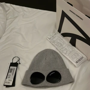 Grå mössa från C.P. Company - Snygg grå ribbad mössa från C.P. Company med inbyggda glasögon. Perfekt för att ge en unik stil till din outfit. Mössan är tillverkad i bomull och har en modern design. Köpt från NK i Stockholm.