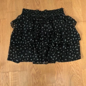 Svart blommig kjol med volanger - Charmig svart kjol med ett blommigt mönster i vitt och grönt. Kjolen har flera lager av volanger som ger en lekfull look. Perfekt för att ge en romantisk touch till din outfit.