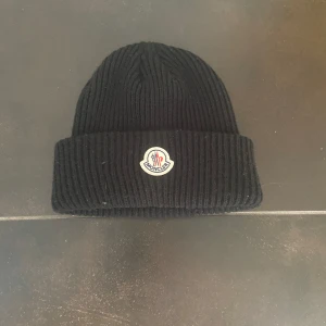 Svart ribbad mössa från Moncler - Snygg svart ribbad mössa från Moncler med broderad logotyp framtill. Perfekt för kyliga dagar och ger en stilren look. Tillverkad i ett mjukt och bekvämt material.
