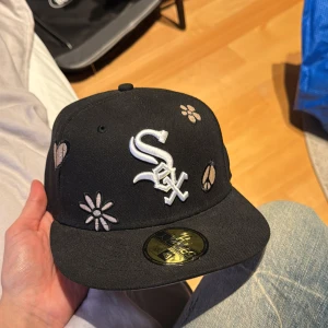 Svart keps med broderade detaljer New era - Snygg svart keps med broderade blommor, hjärtan och fredssymboler. Kepsen har en vit logotyp framtill och är perfekt för en cool och avslappnad stil. (55,8cm)