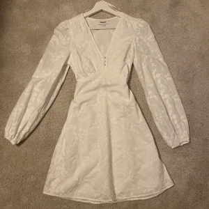 Vit klänning  - Elegant vit klänning med v-ringning och långa ärmar. Klänningen har ett subtilt blommigt mönster och dekorativa knappar framtill. Perfekt för en stilren look. Klänningen är endast använd en gång!❤️