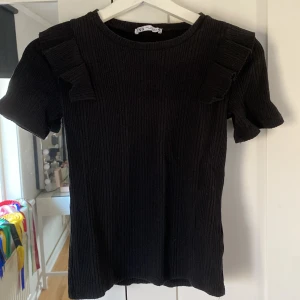 Svart ribbad topp från Zara - Snygg svart ribbad topp från Zara med korta ärmar och dekorativa volanger på axlarna. Perfekt för en stilren look. Passar bra till både jeans och kjol.