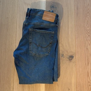 Blå jeans från Jack & Jones - Snygga blå jeans från Jack & Jones med klassisk femficksdesign och en läderpatch på baksidan. De har en rak passform och är perfekta för en avslappnad stil. Jeansen har en knappgylf och är tillverkade i slitstarkt denim. De är i modellen Slim Glenn. 