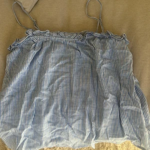 Blå- och vitrandig linnetopp - Säljer en söt blå- och vitrandig linnetopp med smala axelband och volangdetaljer vid halsringningen. Perfekt för varma sommardagar. Toppen har en luftig och bekväm passform.