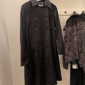 Svart trenchcoat - Snygg svart trenchcoat med dubbelknäppning och klassisk krage. Perfekt för höst och vår, med en stilren design som passar till många tillfällen. Långärmad och med en elegant passform. Två fickor framtill