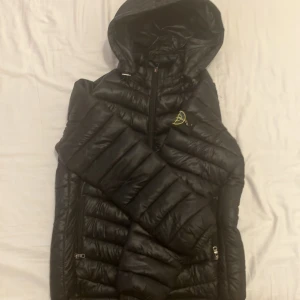 Svart dunjacka från Stone Island - Säljer en svart dunjacka från Stone Island med huva och dragkedja. Jackan har en klassisk pufferdesign och är perfekt för kyligare väder. Märkets ikoniska logga finns på ärmen. Perfekt för dig som vill ha stil och funktion i ett.