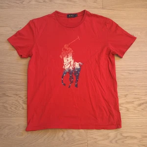 Röd t-shirt från Polo Ralph lauren - Säljer en snygg röd t-shirt från Polo med ett stort tryck av en polospelare på framsidan. T-shirten är tillverkad i 100% bomull och har en klassisk passform med korta ärmar. Perfekt för en avslappnad stil. Den är liten i storlek så skulle säga att den passar M eller S bättre. Säljer den för att jag håller på och rensar ut saker som inte passar längre