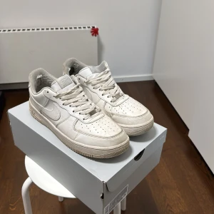 Vita Nike Air Force sneakers - Snygga vita Nike Air Force sneakers med klassisk design. Skorna har en robust sula och ikonisk Nike-logga på sidan och hälen. Perfekta för en stilren look.