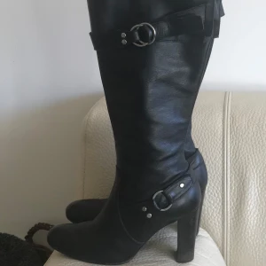 Svarta stövlar med klack - Snygga svarta stövlar i skinn med klack och dekorativa spännen. Perfekta för en stilren look. De har en elegant design som passar till många olika outfits.