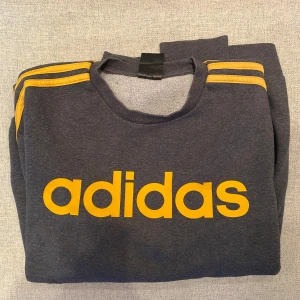 Grå tröja från Adidas med gula detaljer - Säljer en snygg grå tröja från Adidas med gula ränder på ärmarna och stort Adidas-tryck på bröstet. Perfekt för en sportig look eller avslappnad stil. Tröjan har långa ärmar och är gjord i ett mjukt material.