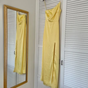 Gul långklänning med slits - Snygg gul långklänning i satin med en elegant slits framtill. Klänningen har en one-shoulder design. Storlek XS och passar mig som är 162 cm. Använd endast en gång. Den är lite insydd i ryggen för att passa bättre.