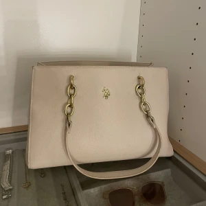 Beige handväska med gulddetaljer - Säljer en jätte fin U.S POLO ASSN väska. Det syns att väskan är använd på kedjan som håller bandet men inget man tänker på. Skriv för fler bilder, priset kan diskuteras!💗