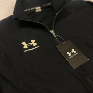 Svart jacka från Under Armour - Säljer en svart jacka från Under Armour med dragkedja och långärmad design. Perfekt för träning eller vardagsbruk. Jackan har en diskret logga på bröstet och är tillverkad i ett syntetmaterial som ger en bekväm passform.