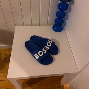 Blå boss tofflor  - Snygga blå sandaler med vit text på remmen. Perfekta för en avslappnad stil och enkla att glida i. De har en modern design och är bekväma för sommarens alla äventyr.