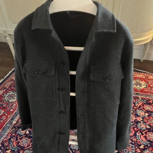 Mörkgrå ulljacka - Snygg mörkgrå ulljacka med knappar framtill och två bröstfickor med lock. Jackan har en klassisk krage och långa ärmar, perfekt för kyligare dagar.