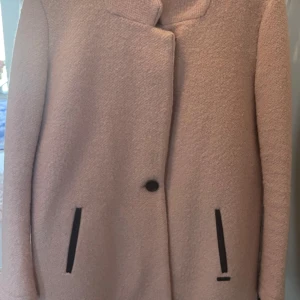 Rosa kappa från Maison Scotch - Säljer en stilren rosa kappa från Maison Scotch. Den har en enkel knäppning med en knapp och svarta detaljer vid fickorna. Perfekt för att hålla stilen under kyligare dagar.
