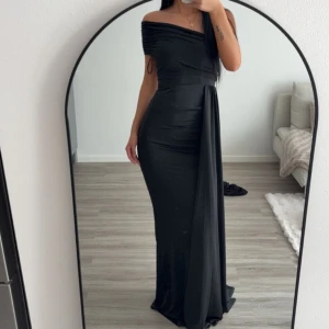 Svart offshoulder långklänning - Elegant svart långklänning med offshoulder och draperad detalj över ena axeln. Klänningen har figurnära passform och ett snyggt fallande tyg framtill. Perfekt för dig som vill sticka ut med en stilren look. Xs passar S. aldrig använt 