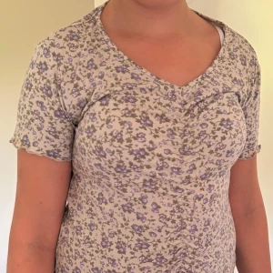 Blommig topp med v-ringning - Supersöt topp med små lila blommor på beige botten. V-ringad hals och korta ärmar med vågig kant. Toppen har rynkade detaljer framtill som ger en snygg passform. Perfekt för dig som gillar romantisk och trendig stil.