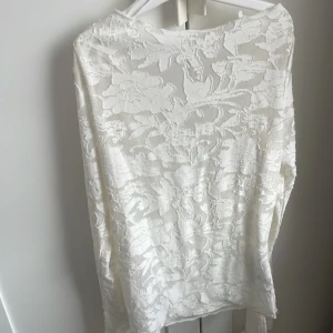 Vit blommig spetsblus med drapering -  Vit långärmad blus i transparent spets med blommigt mönster och en rak hals.