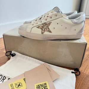 Golden Goose sneakers med glitterstjärna - Snygga sneakers från Golden Goose i vitt skinn med beige mockadetaljer och en glittrig stjärna i guld på sidan. Skorna har rund tå, platt sula och klassisk snörning. Perfekta för dig som vill sticka ut med en cool detalj.