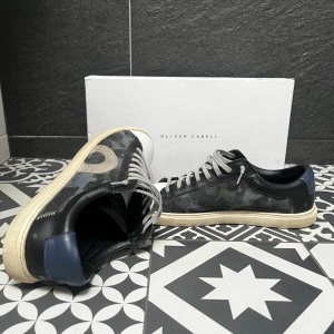 Oliver Cabell Skor - Snygga sneakers från Oliver Cabell med svart och grått camouflagemönster, vit tå och beige sula. Skorna har grå snörning, blå detalj på hälen och är tillverkade i skinn och mocka. Perfekta nu för sommaren med dessa stilrena skor. Pris kan diskuterad vid snabb affär!