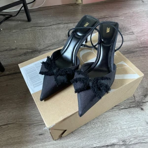 Svarta pumps med rosett från Zara - Stilrena svarta pumps från Zara med spetsig tå och smal klack. Skorna har dekorativa rosetter i fransigt tyg framtill och tunna remmar runt hälen. Snygg satinliknande finish som ger en lyxig känsla. Ungefär 7 cm klack 