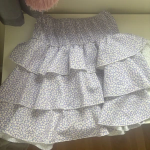 Ljuslila blommig volangkjol - Supersöt kort kjol i ljuslila med små vita blommor och flera volanger. Resår i midjan för skön passform. Perfekt för vår och sommar när du vill ha en lekfull och trendig look.