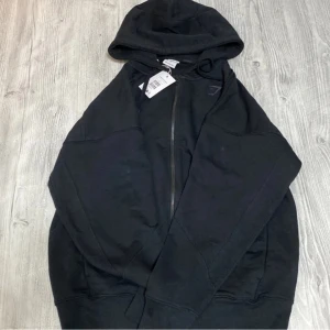 Svart Gymshark hoodie med dragkedja - Svart hoodie från Gymshark i storlek S med hel dragkedja framtill och stor logga på ryggen. Tröjan har huva med snörning och två fickor. Perfekt för dig som gillar sportig och avslappnad stil.