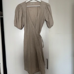 Beige omlottklänning från Gina Tricot - Supersnygg beige omlottklänning från Gina Tricot i linneblandning. Klänningen har puffiga korta ärmar, knytband i midjan och v-ringning. Perfekt för dig som gillar en avslappnad men trendig stil. Använd en gång.