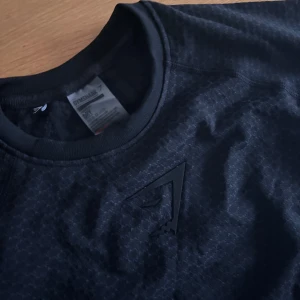 Svart Gymshark ONYX V3 - Svart t-shirt från Gymshark i funktionsmaterial med diskret mönstrad struktur och tryckt logga på bröstet. Modellen är kortärmad och har en sportig vibe, perfekt för träning. Materialet är lätt och andas, vilket gör den skön att ha på sig under gympass. Priset är diskuterbart vid en snabb och lätt affär.