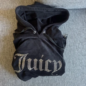 Juicy Couture svart velourjacka S - Svart velourjacka från Juicy Couture med huva och snörning. Stor Juicy-logga i glittrande stenar på bröstet. Klassisk 2000-talsvibe, perfekt för dig som vill ha en ikonisk och mjuk hoodie med bling. Snygg och bekväm att slänga på sig när du vill sticka ut. (Metal grejerna på snöret har lossnat)
