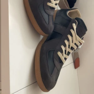 Svarta sneakers från Maison Margiela -  INTRESSEKOLL ‼️.                                    Snygga bruna sneakers från Maison Margiela med beige snörning och klassisk gummisula. Skorna har en mix av mocka och skinn, vilket ger en stilren och tidlös look. Perfekta för dig som gillar streetstyle och vill ha något unikt i garderoben.
