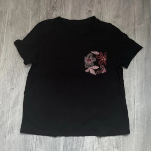 Svart t-shirt med blommig ficka - Svart t-shirt med korta ärmar och en snygg bröstficka med broderade blommor i rosa, rött och vitt. Fickan ger en cool kontrast och extra detalj till den annars enkla t-shirten. Loose fit, storlek Small. Lappar avklippta.