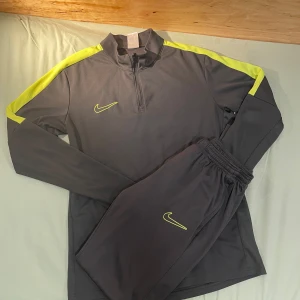 Nike Dri-Fit grå träningsöverdel M - Nike Dri-Fit långärmad träningsöverdel i mörkgrått med neongula detaljer längs axlarna och halv dragkedja framtill. Perfekt för fotboll eller träning, tillverkad i lätt och snabbtorkande polyester. Snygg och sportig look med klassisk Nike-logga på bröstet.