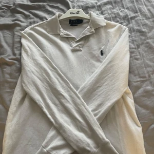 Vit långärmad pikétröja Polo Ralph Lauren - Vit långärmad pikétröja från Polo Ralph Lauren i klassisk modell med krage och knappar framtill. Tröjan har det ikoniska broderade logotypen på bröstet och är tillverkad i bomull. Perfekt för en clean och stilren look.