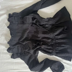 Svart blus med volangdetaljer - Svart blus med långa ärmar och snygga volangdetaljer längs axlar och bröst. Blusen har en lätt skrynklig struktur och rundad halsringning med volangkant. Perfekt för dig som gillar en trendig och feminin look.